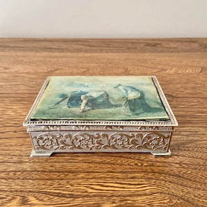 以下が含まれることがあります： 縁に装飾的な花柄が施された銀色の金属製の小物入れ。箱の上面には、畑で働く人々のヴィンテージ風の画像が描かれています。
