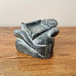 May include: A black stone sculpture of a person in a seated position, with a square-shaped indentation in the top, likely used as an ashtray.