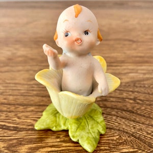 May include: A porcelain figurine of a baby sitting in a yellow flower. The baby has a light pink body, orange hair, and brown eyes. The flower has green leaves.