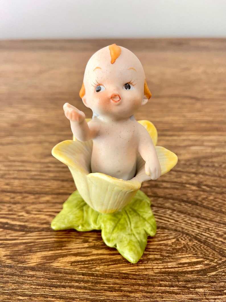May include: A porcelain figurine of a baby sitting in a yellow flower. The baby has a light pink body, orange hair, and brown eyes. The flower has green leaves.