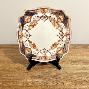 Peut inclure: Assiette carrée en céramique avec un motif floral. L'assiette présente un centre blanc avec une bordure de fleurs orange, jaunes et rouges. Le bord extérieur a un motif bleu marine et or. L'assiette est présentée sur un support noir.