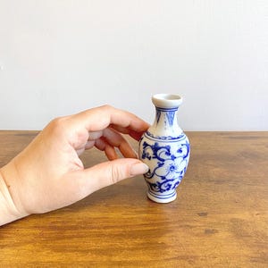 Può includere: Un piccolo vaso in ceramica bianca con motivi floreali blu. Il vaso ha un collo stretto e un corpo bulboso. Il design blu include fiori e motivi a spirale. Il vaso è tenuto contro una superficie di legno.