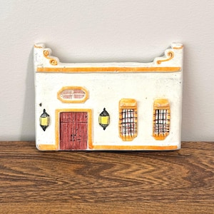 May include: A rectangular ceramic wall plaque shaped like a building facade with a white exterior, a dark red door, two barred windows, and two yellow lanterns. The structure features orange trim accents and a decorative top edge with scroll details.