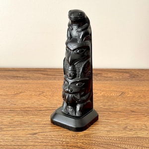 May include: A black totem pole sculpture with carved animal figures. The sculpture features a frog, an eagle, and a bear. The totem pole is displayed on a wooden surface.