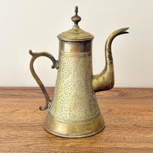 May include: A brass coffee pot featuring an intricate engraved floral pattern across its conical body. The vessel includes a curved handle, a long tapered spout, and a removable lid topped with a small finial. The metal shows a weathered patina.