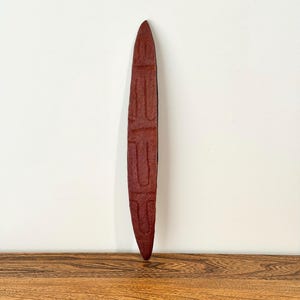 May include: A long, slender, pointed wooden object with a deep reddish-brown finish. The surface features a series of carved, geometric, and linear patterns that create a textured appearance across the entire length of the elongated form.