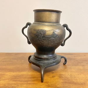 May include: Antique bronze-colored vase with a detailed landscape scene. The vase has two curved handles and sits on a tripod base. The landscape features a building, trees, and mountains. The vase is approximately 8 inches tall.