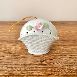 May include: White ceramic ornament shaped like a basket with a pink rose and green leaves. The ornament has a brown string for hanging.