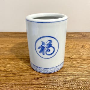 May include: White ceramic cup with a blue circular design and Chinese characters. The cup has a blue wave pattern around the base and a blue rim. The cup is cylindrical and sits on a wooden surface.