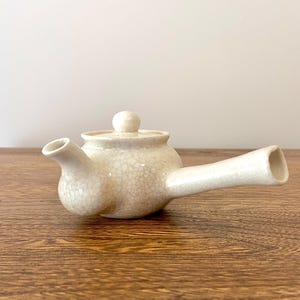 Puede incluir: Una tetera de cerámica blanca con un esmalte craquelado y un pico largo. La tetera tiene un cuerpo redondo y una tapa pequeña.