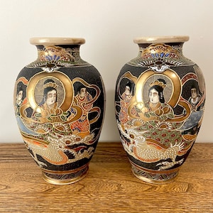 May include: Two matching antique Japanese vases. Each vase features a black background with gold accents and detailed illustrations of figures and dragons. The vases have a rounded shape with a narrow neck and a gold rim.