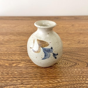 Può includere: Un piccolo vaso in ceramica bianca con una glassa macchiettata e un motivo floreale marrone e blu.