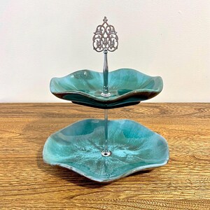 Puede incluir: Fuente de dos niveles con platos turquesa con bordes ondulados. Los platos están conectados por una varilla central plateada con un asa ornamentada. La fuente está sobre una superficie de madera.