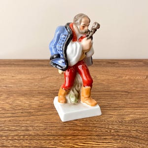 Puede incluir: Figura de cerámica de un hombre con vestimenta tradicional, con chaqueta azul, camisa blanca y pantalón rojo. La figura sostiene un instrumento musical y se encuentra sobre una base blanca. Las botas son color canela.