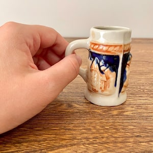 May include: A small white ceramic beer stein with a blue and orange design. The stein has a handle and a raised design of a building.