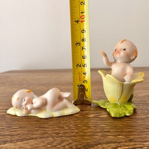 May include: Two porcelain figurines of babies. One is lying down on a yellow and white blanket, the other is sitting in a yellow flower with green leaves. The figurines are approximately 3 inches tall.