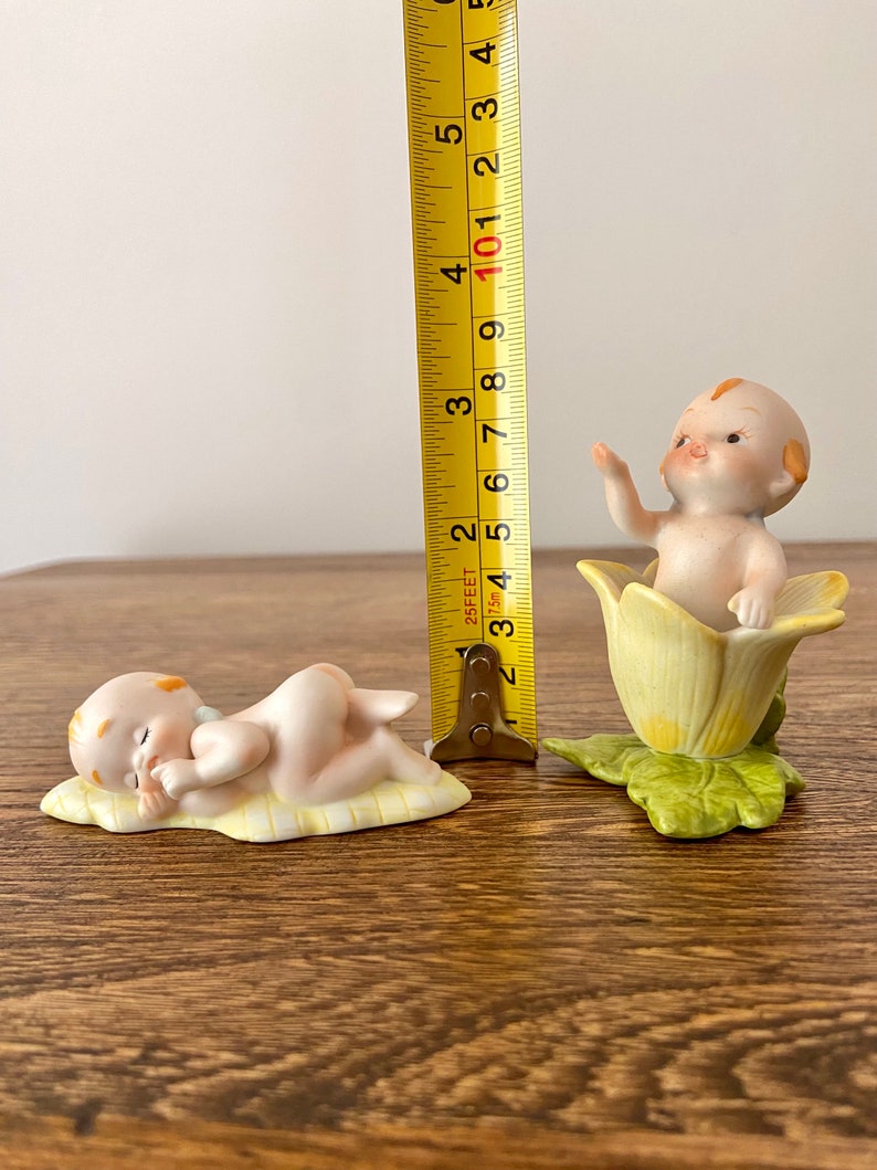 May include: Two porcelain figurines of babies. One is lying down on a yellow and white blanket, the other is sitting in a yellow flower with green leaves. The figurines are approximately 3 inches tall.