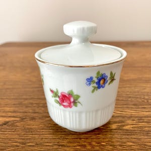May include: White porcelain sugar bowl with a lid and a gold trim. The bowl is decorated with a floral pattern of pink, blue, and green flowers.