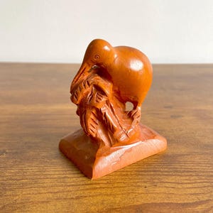 Puede incluir: Una escultura de madera de un kiwi, tallada en un tono marrón cálido. El ave está posada sobre una base rectangular con follaje tallado. La escultura es detallada, mostrando el pico y el cuerpo del ave.