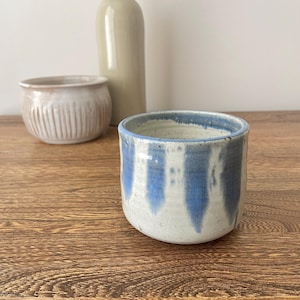 Könnte beinhalten: Ein weißer Keramiktopf mit blauen vertikalen Streifen. Der Topf steht auf einer Holzoberfläche.