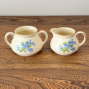Peut inclure: Un sucrier en céramique crème à deux anses et un pot à lait assorti, tous deux ornés d'un motif floral bleu et d'un liseré doré sur les bords et les anses, présentés comme un ensemble pour servir le café ou le thé.