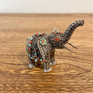 May include: A detailed silver elephant figurine with turquoise and coral accents. The elephant has intricate filigree detailing and is posed with its trunk raised. The figurine is displayed on a wooden surface.