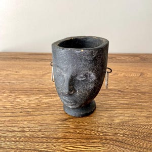 May include: A black ceramic planter shaped like a human face with a textured surface. The planter has two small holes on either side of the face, which could be used for earrings.