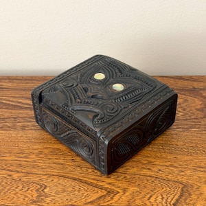 Puede incluir: Una caja de madera cuadrada de color marrón oscuro con intrincados diseños tallados. La tapa presenta un motivo central con dos incrustaciones ovaladas iridiscentes. Los lados de la caja también muestran tallados detallados, mostrando una estética artesanal.