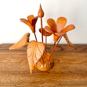 May include: A wooden floral sculpture featuring carved orange flowers, buds, and leaves. The handcrafted piece is set in a round base and sits on a wooden surface. The sculpture is a decorative item.