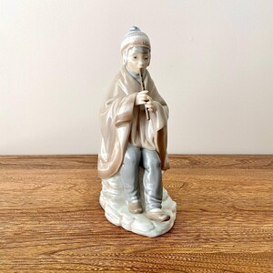 Può includere: Una statuetta in porcellana di una persona che indossa un mantello marrone con cappuccio e un cappello bianco, che suona il flauto mentre è seduta su uno scoglio.