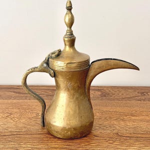 May include: A brass dallah coffee pot featuring a wide base, a long curved spout, and a tall, pointed lid finial. The metal surface shows a weathered patina with a textured handle and a small stamped mark on the side of the main body.