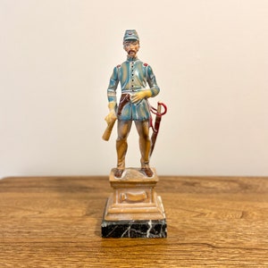 May include: A painted figurine of a soldier in a blue uniform with a red sash and a white hat. The soldier is holding a rolled up scroll in one hand and a sword in the other. The figurine is standing on a marble base.