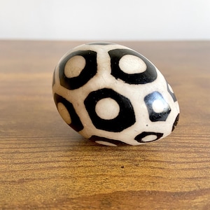 May include: A decorative stone with a distinctive pattern. The stone is oval-shaped, featuring a cream-coloured base adorned with a repeating design of black circles and curved shapes. It rests on a wooden surface. The stone could be a bead or amulet.