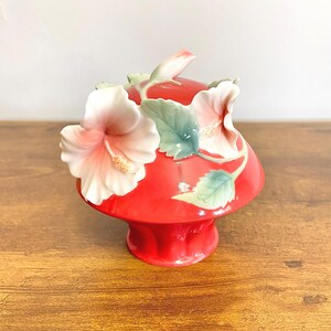 May include: A red ceramic decorative container with a lid, decorated with detailed hibiscus flowers in shades of pink and white, and green leaves. The container has a pedestal base and is set on a wooden surface.