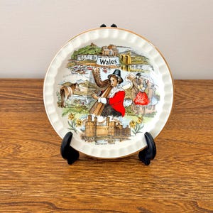 May include: A round ceramic decorative plate with a scalloped edge and gold trim, featuring a colourful illustration of Wales. The design includes a harpist, a Welsh pony, Caernarvon Castle, and the text Wales, Tenby Harbour, and Menai Bridge.