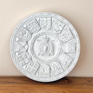 May include: A white, circular commemorative plaque with the text "Centennial 1873-1973 Royal Canadian Mounted Police." The plaque has a detailed relief design with various emblems and a central crest. The plaque is on a wooden surface.