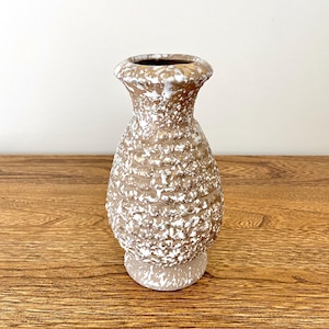 May include: A textured, brown and white ceramic vase with a bulbous body and a narrow neck. The vase has a speckled, rough surface and a small, round base. The interior of the vase is dark. The vase is displayed on a wooden surface.