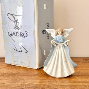 May include: A porcelain angel figurine with a flowing white dress and light blue accents. The angel has detailed wings and holds a scroll. The box behind the figurine has the word "LLADRÓ" printed on it.