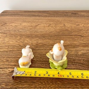 May include: Two porcelain figurines of children in a flower pot and lying down. The figurines are painted in pastel colors and have a vintage look.