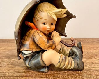 May include: A vintage porcelain figurine of a child seated under a brown umbrella. The child wears a plaid shirt, dark shorts, and tall socks. The umbrella is brown with a curved handle, and the figurine has a detailed, hand-painted appearance.