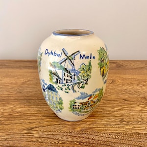 Puede incluir: Un jarrón de cerámica blanca con un diseño pintado en azul y verde de un molino de viento y edificios. El jarrón tiene las palabras "Dybbøl" y "Mølle" escritas en él.