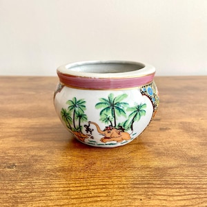 Puede incluir: Un pequeño macetero de cerámica blanca con un borde rosa y un diseño colorido de elefantes y palmeras.