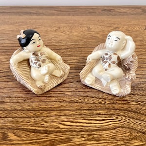 May include: Two small figurines of children sitting in chairs. The figurines are made of white ceramic and have painted details. The girl figurine has black hair and a red bow. The boy figurine has black hair and is wearing a brown and white patterned shirt. The chairs are made of brown ceramic and have a woven pattern.