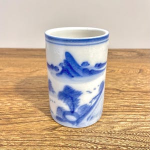 Könnte beinhalten: Eine zylindrische Keramiktasse mit weißem Grund und blauem Landschaftsdesign. Das Design zeigt Berge, Bäume und andere natürliche Elemente. Die Tasse hat ein schlichtes, elegantes Design und eignet sich zum Trinken.