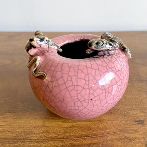 Może przedstawiać: Różowa ceramiczna miska z pękniętą glazurą, z dwiema figurkami żab na obrzeżu. Miska ma ciemne wnętrze i stoi na drewnianej powierzchni. Żaby mają zielone i brązowe akcenty.