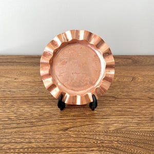 May include: A small, copper-colored ashtray with a scalloped edge. The ashtray features an engraved design of a map of the Hawaiian Islands with the text "Hawaiian Islands" and "1957".