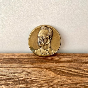 Puede incluir: Una moneda de metal de color dorado con una imagen en relieve de un rostro masculino. El rostro del hombre está parcialmente oscurecido por una imagen esquelética.