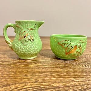Op de afbeelding: Een groene keramische roomkan en suikerpot set met bladgoud accenten. De roomkan heeft een handvat en een getextureerd oppervlak. De suikerpot is rond en heeft een getextureerd oppervlak.