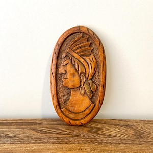 May include: A wooden wall hanging featuring a carved profile of a person wearing a headdress. The carving is oval shaped and has a brown wood finish.