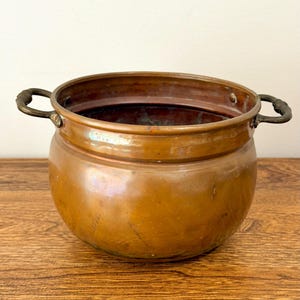 May include: A copper pot with two brass handles. The pot has a rounded body and a wide opening. The copper has a warm, reddish-brown color and a slightly reflective surface. The pot is sitting on a wooden surface.